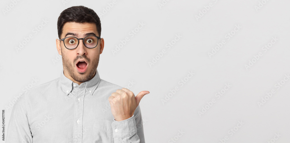 Naklejka premium Banner of surprised young man in shirt pointing with thumb to the right, isolated on gray background with copy space