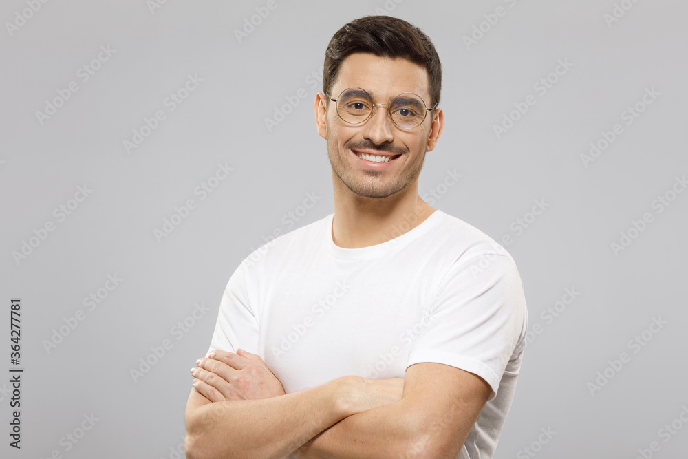 Portrait of handsome smiling guy dressed in white t-shirt, wearing round eyeglasses, holding arms crossed, ready to help and consult, isolated on gray background Portrait of handsome smiling guy dressed in white t-shirt, wearing round eyeglasses, holding arms crossed, ready to help and consult, isolated on gray background