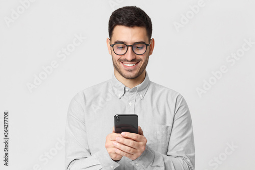 Young man wearing gray shirt and glasses, holding his smartphone and exchanging messages with friends with smile, isolated on studio background