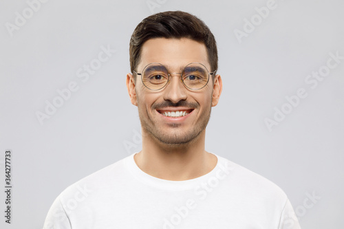 Close-up portrait of young smiling handsome man wearing white t-shirt and glasses, feeling optimistic and confident, isolated on gray background © Damir Khabirov
