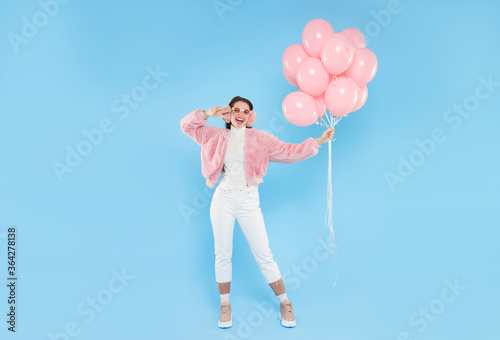 Full length portrait of young happy female wearing pink fluffy bomber and earwarmers, standing with pink birthday balloons, isolated on blue background