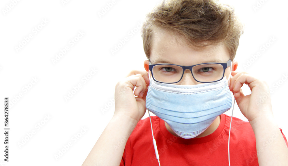 Portrait of a boy in a surgical medical blue mask against covid-19 coronavirus. A teenager in white wired headphones, glasses, and a red t-shirt listens to music on a white background. Copyspace.