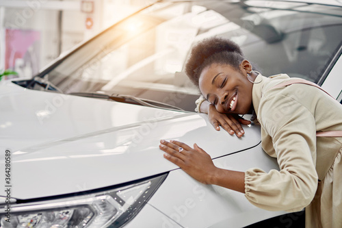 smiling black lady hugs her new auto in cars showroom. young woman fulfill her dream, lies on car