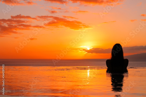 A girl stands with her back in the pool and looks at the setting sun in the ocean.