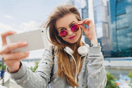 young hipster woman having fun in street, wearing pink sunglasses, student on vacation, spring summer urban style, taking selfie pisture on smartphone photo