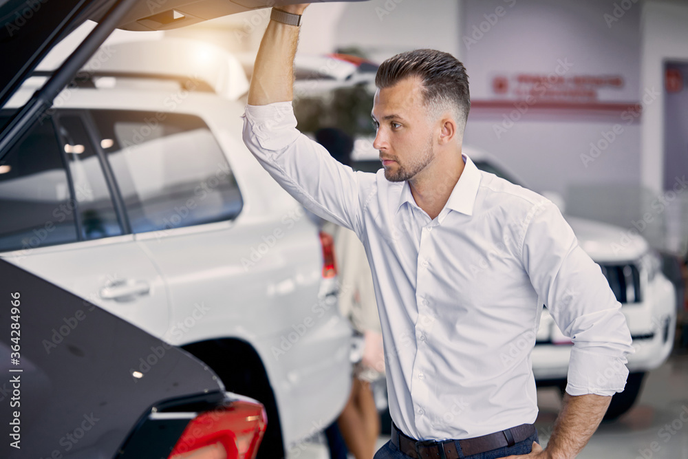 handsome caucasian guy examine the trunk of car in dealership , he looks inside of car from back and think buy it or not handsome caucasian guy examine the trunk of car in dealership , he looks inside of car from back and think buy it or not