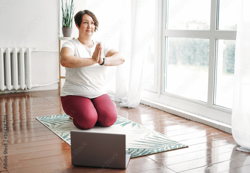 Adult charming brunette woman plus size body positive practice yoga with laptop at the home - obrazy, fototapety, plakaty 