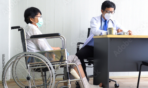 asian doctor wearing mask examines the woman patient who sits in wheelchair because she gets leg injury. healthcare and medical concept