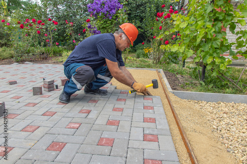 A gloved craftsman lays paving stones in layers. Brick paving slabs for professional use. Laying gray concrete paving slabs in the courtyard of the house on a sandy foundation.
