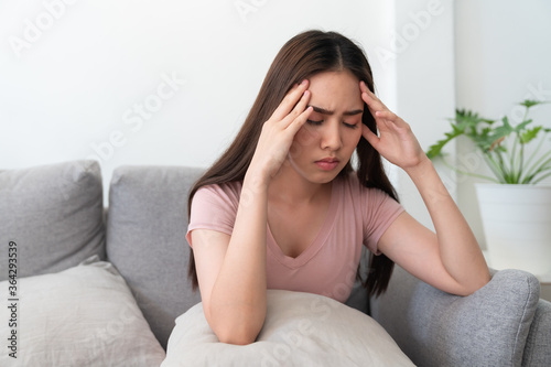 Asian young woman holding her head,she has a headache sitting on sofa couch in living room at home.
