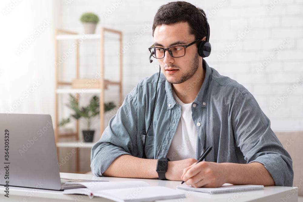 Fototapeta premium Teacher online. Guy in glasses and headset makes notes in notebook and looks at laptop