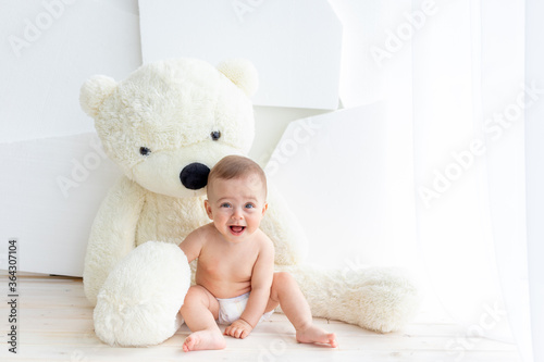A small child a girl of 6 months is sitting with a large soft bear in a bright apartment in diapers © Any Grant