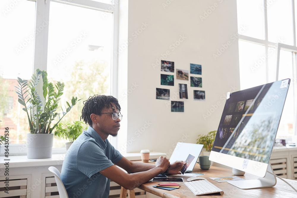 Portrait of young African-American photographer using computer at desk in home office with photo editing software on screen, copy space - obrazy, fototapety, plakaty 