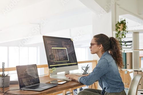 Side view portrait of female IT developer typing on keyboard with black and orange programming code on computer screen and laptop in contemporary office interior, copy space