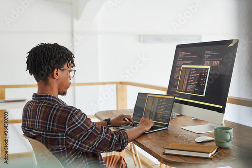 African-American IT developer typing on keyboard with black and orange programming code on computer screen and laptop in contemporary office interior, copy space photo