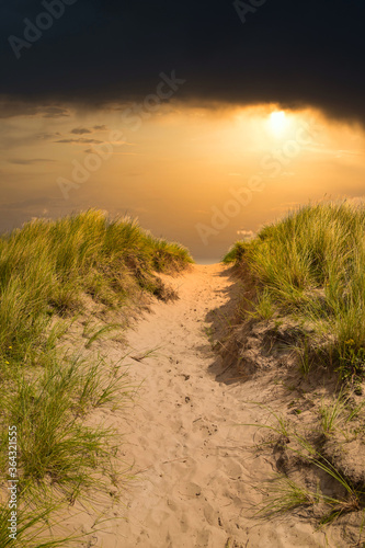 Dramatic seascape of sandy path leading uphill through tall grass on sand dunes. Orange and black sunset sky, golden light on sand and grass. Bamburgh, Northumberland, England.