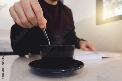 Close up image of a man stirring a coffee cup and reading a book in a cafe.