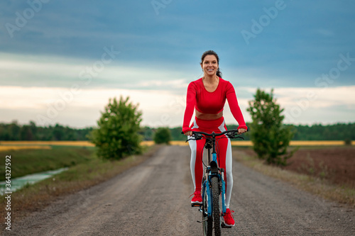 Beautiful girl in a red sports suit on a bicycle on a sunset background. The concept of a healthy lifestyle, sports training, cardio load. Copy space.