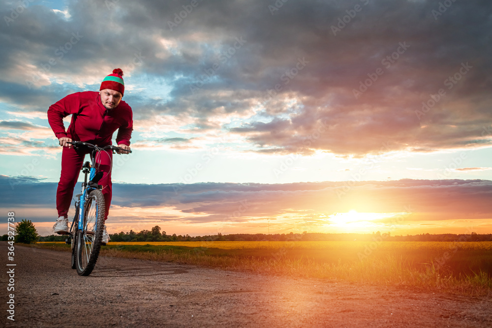 Fototapeta premium Sports training on a bicycle. A man in a tracksuit on a sunset background. The concept of a healthy lifestyle, cardio training, physical activity. Copy space.
