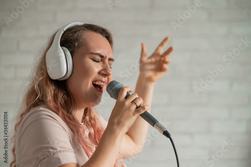 Close-up portrait of a caucasian woman with curly hair singing into a microphone. Beautiful emotional girl in white headphones sings a song in home karaoke and actively gestures against a brick wall.