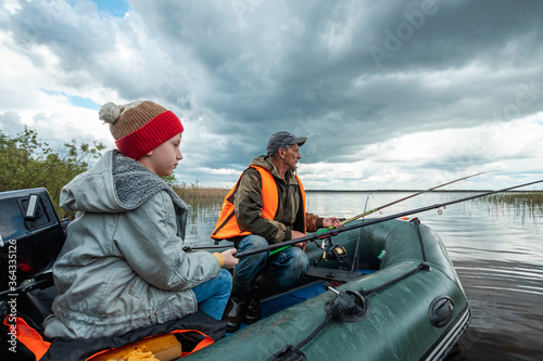 Grandson and grandfather together fish from a boat on the lake. The concept of family, summer vacation, generation. Copy space. © Aliaksandr Marko