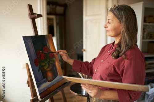 Attractive stylish mature retired female having inspired look holding brush and palette painting flowers on canvas using oil paint, standing in front of easel during art class for beginners