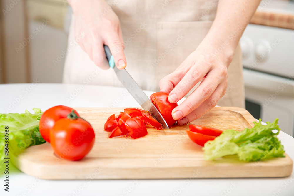 Elegant hands of a young girl cut juicy red tomato into halves on a wooden cutting board. Preparation of ingredients and vegetables. Organic products for diet, nutrition, healthy lifestyle