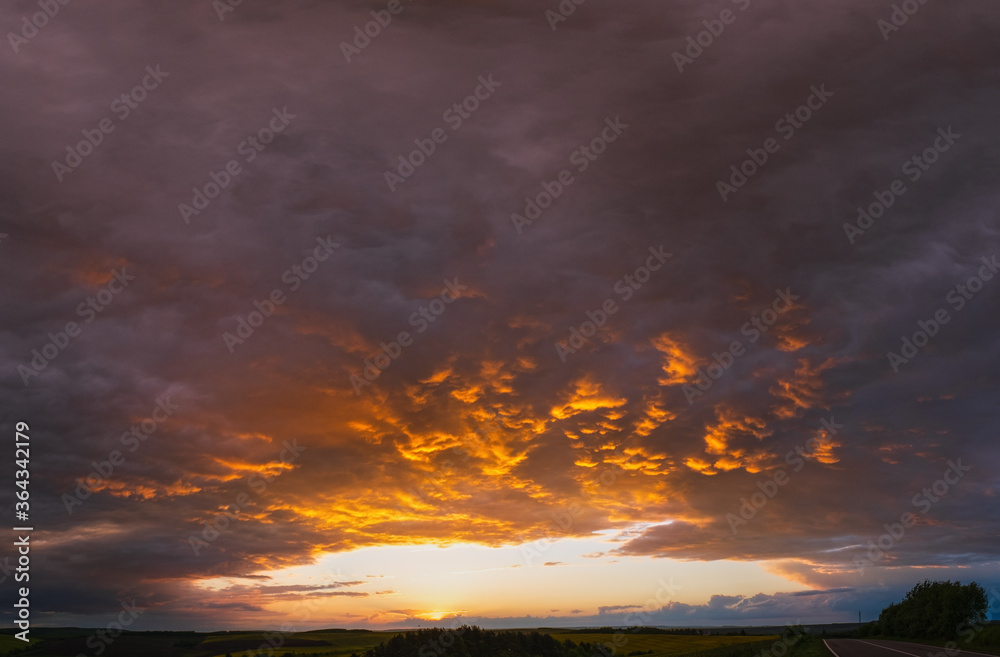 Cloudy sunset evening sky above spring rapeseed yellow fields, road and rural hills. Natural seasonal, weather, climate, countryside beauty concept and background scene. Cloudy sunset evening sky above spring rapeseed yellow fields, road and rural hills. Natural seasonal, weather, climate, countryside beauty concept and background scene.