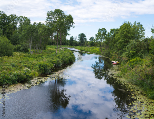 a small, narrow river with the reflection of clouds in the water; there are trees and shrubs on the shores