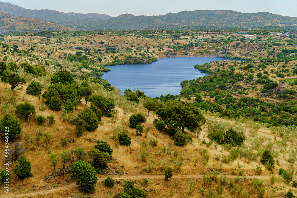blue lake in dry landscape with hills, trees and mountains in the background
