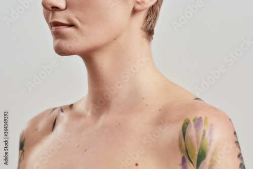 Cropped portrait of a young attractive naked tattooed woman with perfect skin looking away isolated over light background