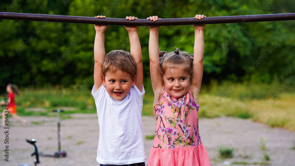 Portrait of funny little girl and boy hanging on horizontal bar in summer park Portrait of funny little girl and boy hanging on horizontal bar in summer park