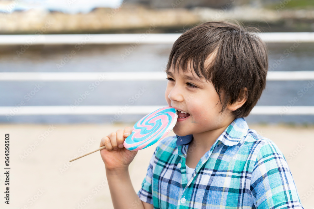 Funny kid eating lollipop, happy little boy holding big sugar candy, Child with smiling face eat sweets, Active childhood enjoying funtime outdoor in sunny day summer Funny kid eating lollipop, happy little boy holding big sugar candy, Child with smiling face eat sweets, Active childhood enjoying funtime outdoor in sunny day summer