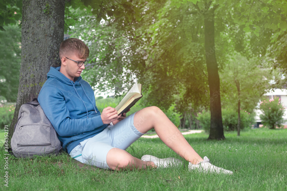 Handsome guy, man, young college or university student or pupil in glasses sitting outdoors in summer park reading book leaning on a tree, learning lessons, preparing for exam in university or college Handsome guy, man, young college or university student or pupil in glasses sitting outdoors in summer park reading book leaning on a tree, learning lessons, preparing for exam in university or college