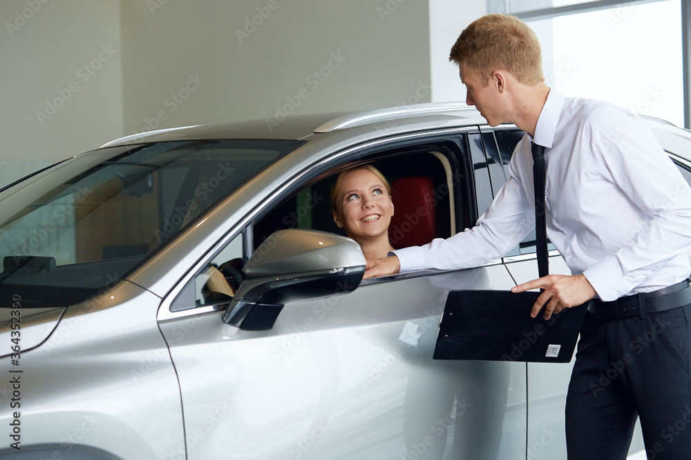 A friendly car dealer shows a young woman a new car explains the pros and cons of a brand new car at the dealership