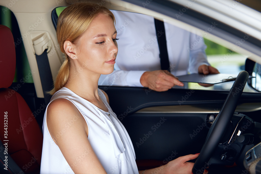 A young woman is sitting in the car enjoying its comfortable compartment listening to the sales agent talk about the new car about the advantages of the new car A young woman is sitting in the car enjoying its comfortable compartment listening to the sales agent talk about the new car about the advantages of the new car