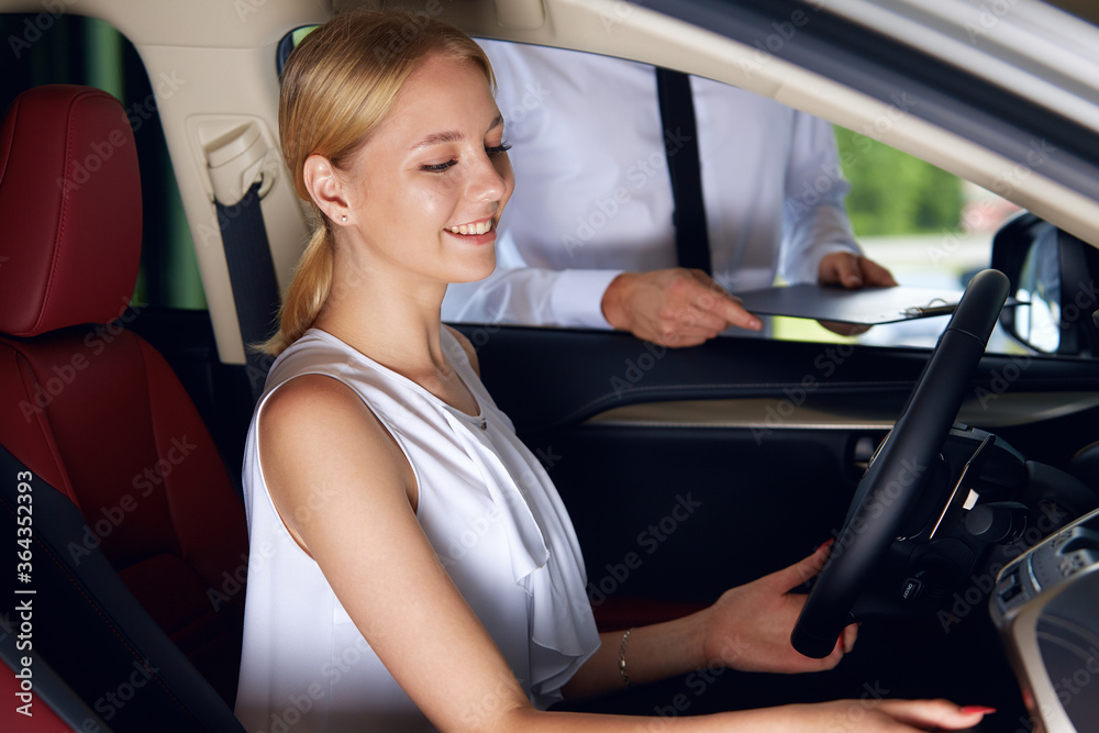 A young woman is sitting in the car enjoying its comfortable compartment listening to the sales agent talk about the new car about the advantages of the new car A young woman is sitting in the car enjoying its comfortable compartment listening to the sales agent talk about the new car about the advantages of the new car