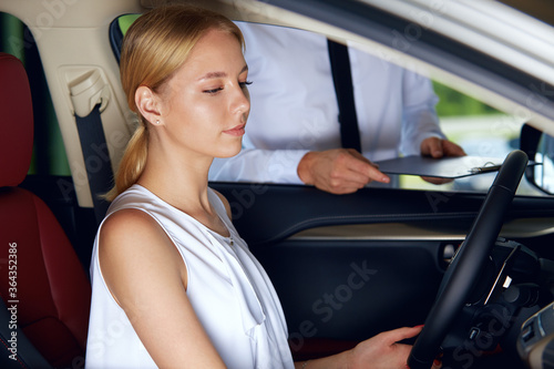 A young woman is sitting in the car enjoying its comfortable compartment listening to the sales agent talk about the new car about the advantages of the new car