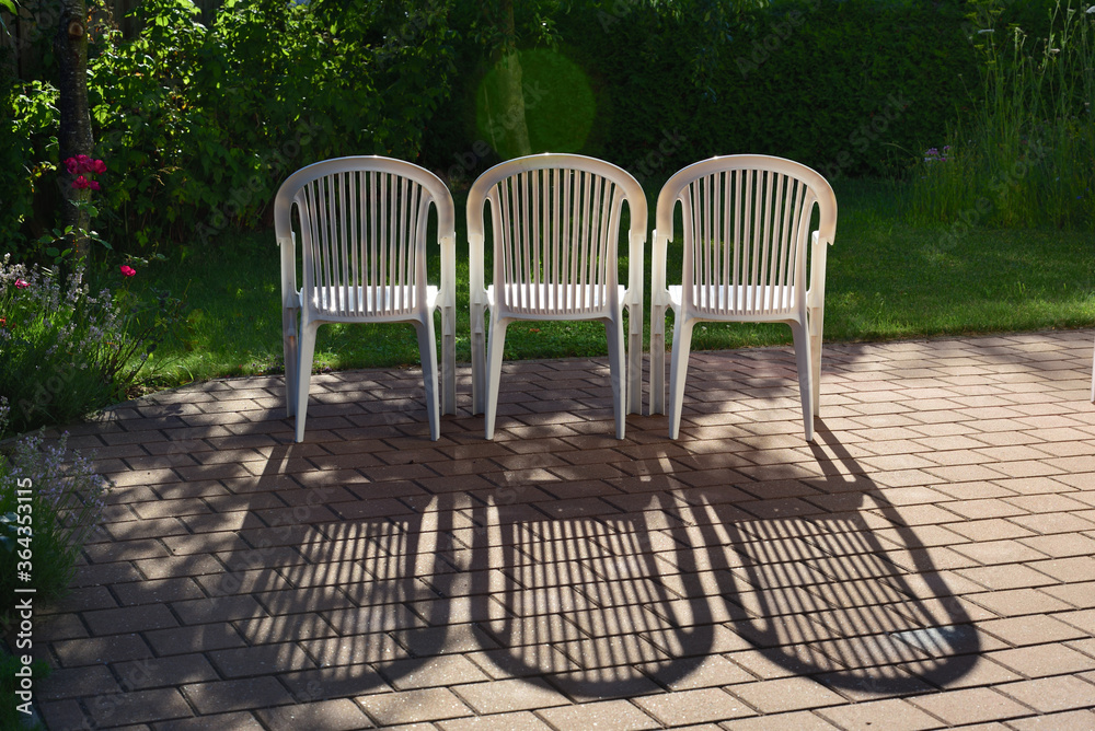 In a romantic garden, three empty white chairs stand side by side and the sun shines through and they cast shadows on the pavement