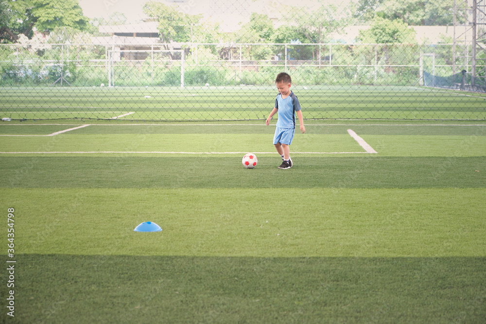 Cute little smiling Asian 4 - 5 years old kindergarten boy, football player in soccer uniform is playing football at Training Session, Soccer Drills for Kids concept, Motion blur at football and hand