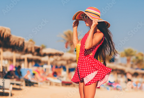 Summer lifestyle portrait of pretty girl walking on the beach of tropical island. Wearing stylish short summer polka-dot dress and straw hat. Holding inflatable yellow beach bag. Tanned fit sexy body.