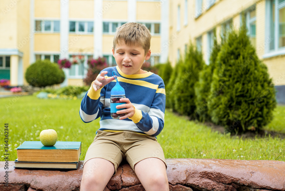 Fototapeta premium Cute schoolboy eating outdoors the school from glass lunch boxe. Food for lunch, lunchboxes with juice, apple and sandwiches, natural fruits.Healthy school breakfast for child.