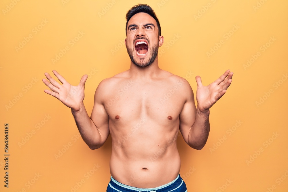 Young handsome man with beard wearing sleeveless t-shirt standing over yellow background crazy and mad shouting and yelling with aggressive expression and arms raised. Frustration concept.