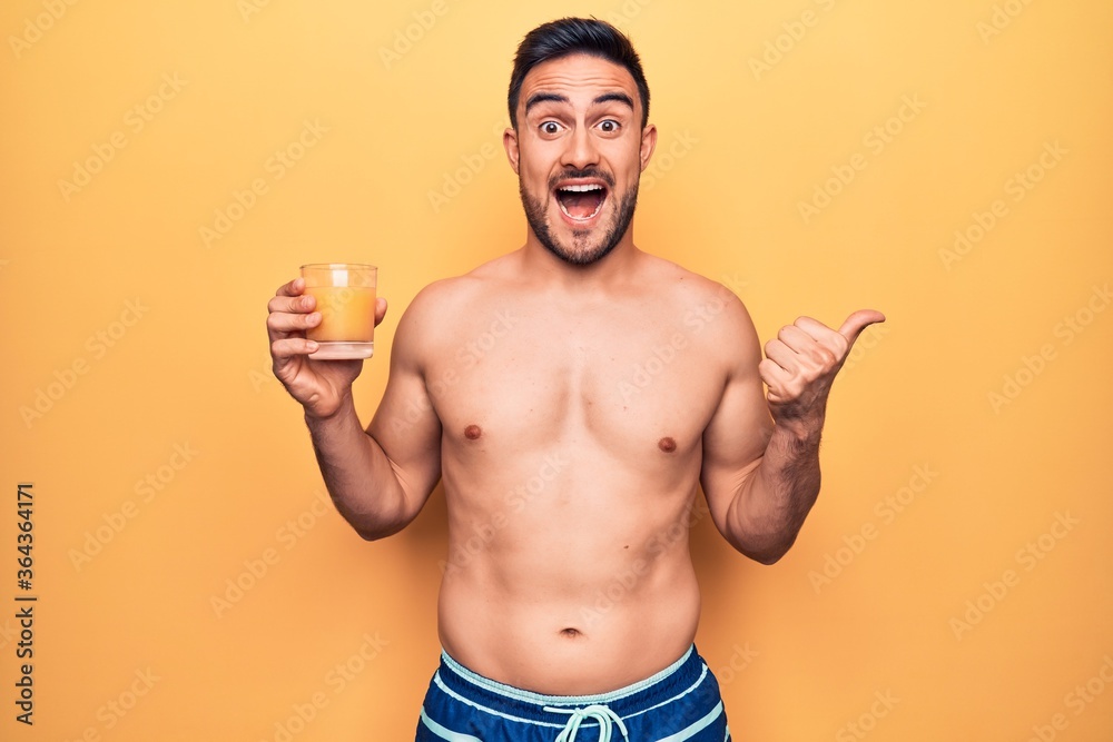 Fototapeta premium Young handsome man with beard on vacation wearing swimwear drinking glass of orange juice pointing thumb up to the side smiling happy with open mouth
