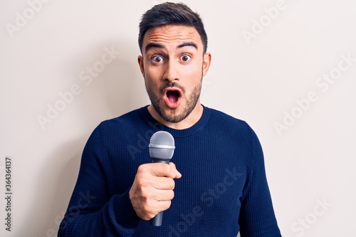 Young handsome singer man with beard singing song using microphone over white background scared and amazed with open mouth for surprise, disbelief face
