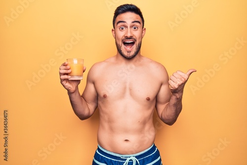 Young handsome man with beard on vacation wearing swimwear drinking glass of orange juice pointing thumb up to the side smiling happy with open mouth