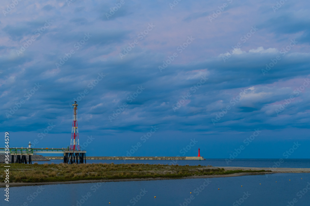 Beautiful seascape of sandbar jutting out into ocean under blue evening sky with red and white tower in front of  red lighthouse on pier.