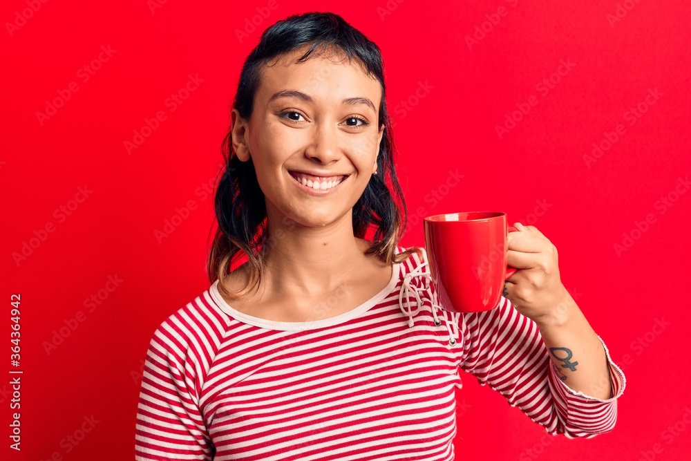 Young woman holding coffee looking positive and happy standing and smiling with a confident smile showing teeth