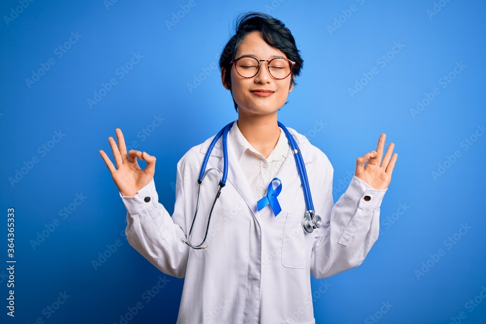Young beautiful asian doctor girl wearing stethoscope and coat with blue cancer ribbon relax and smiling with eyes closed doing meditation gesture with fingers. Yoga concept. Young beautiful asian doctor girl wearing stethoscope and coat with blue cancer ribbon relax and smiling with eyes closed doing meditation gesture with fingers. Yoga concept.