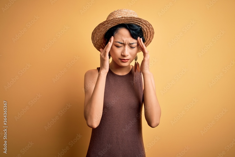 Young beautiful asian girl wearing casual t-shirt and hat over isolated yellow background with hand on head for pain in head because stress. Suffering migraine. Young beautiful asian girl wearing casual t-shirt and hat over isolated yellow background with hand on head for pain in head because stress. Suffering migraine.